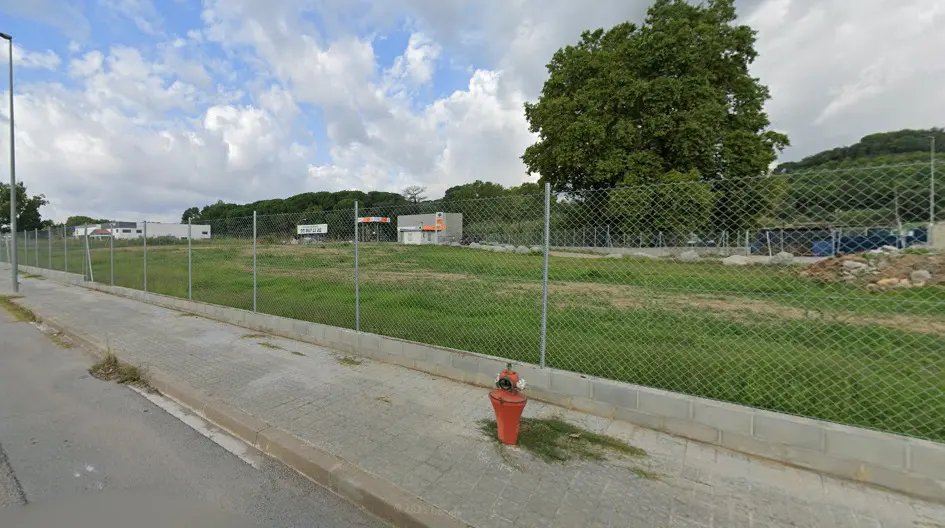 A vacant lot with green grass is enclosed by a chain-link fence. A red fire hydrant stands on the sidewalk in front of the lot.