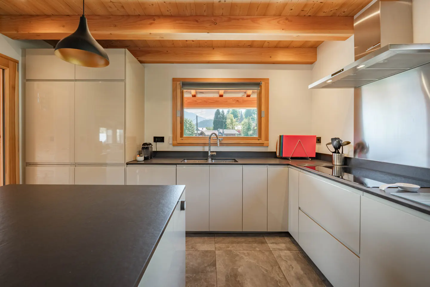 Modern kitchen with white cabinets, dark countertops, and wood beam ceiling. A window overlooks a mountain view.