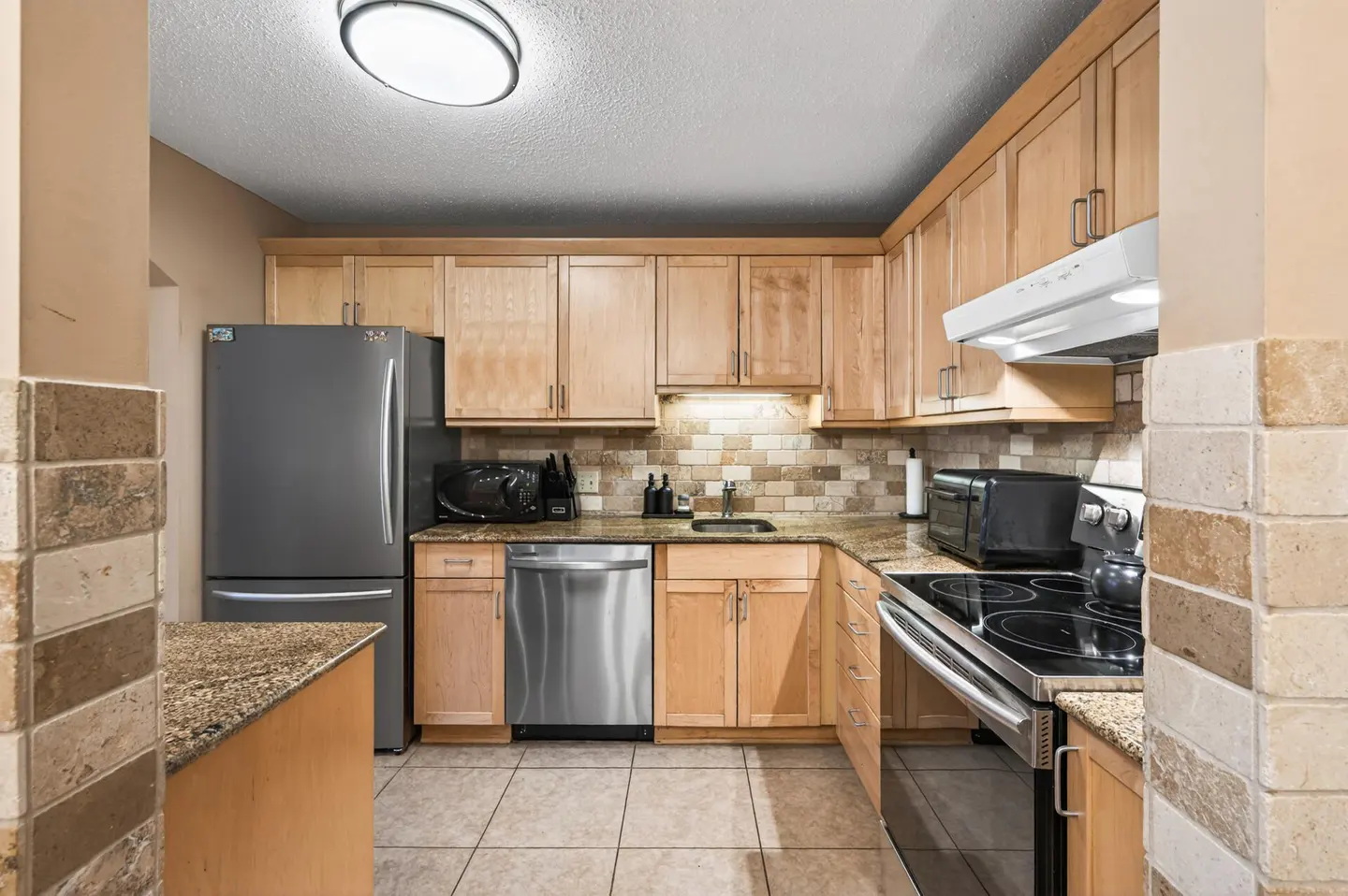 A kitchen with light wood cabinets, stainless steel appliances, and granite countertops. A gray refrigerator is on the left.