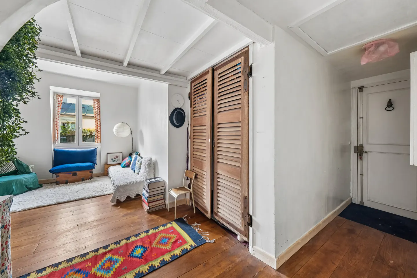 Bright living room with hardwood floors, white walls, and a window. A colorful rug leads to a seating area with a blue couch and a white daybed. A pair of wooden shutter doors are on the right.