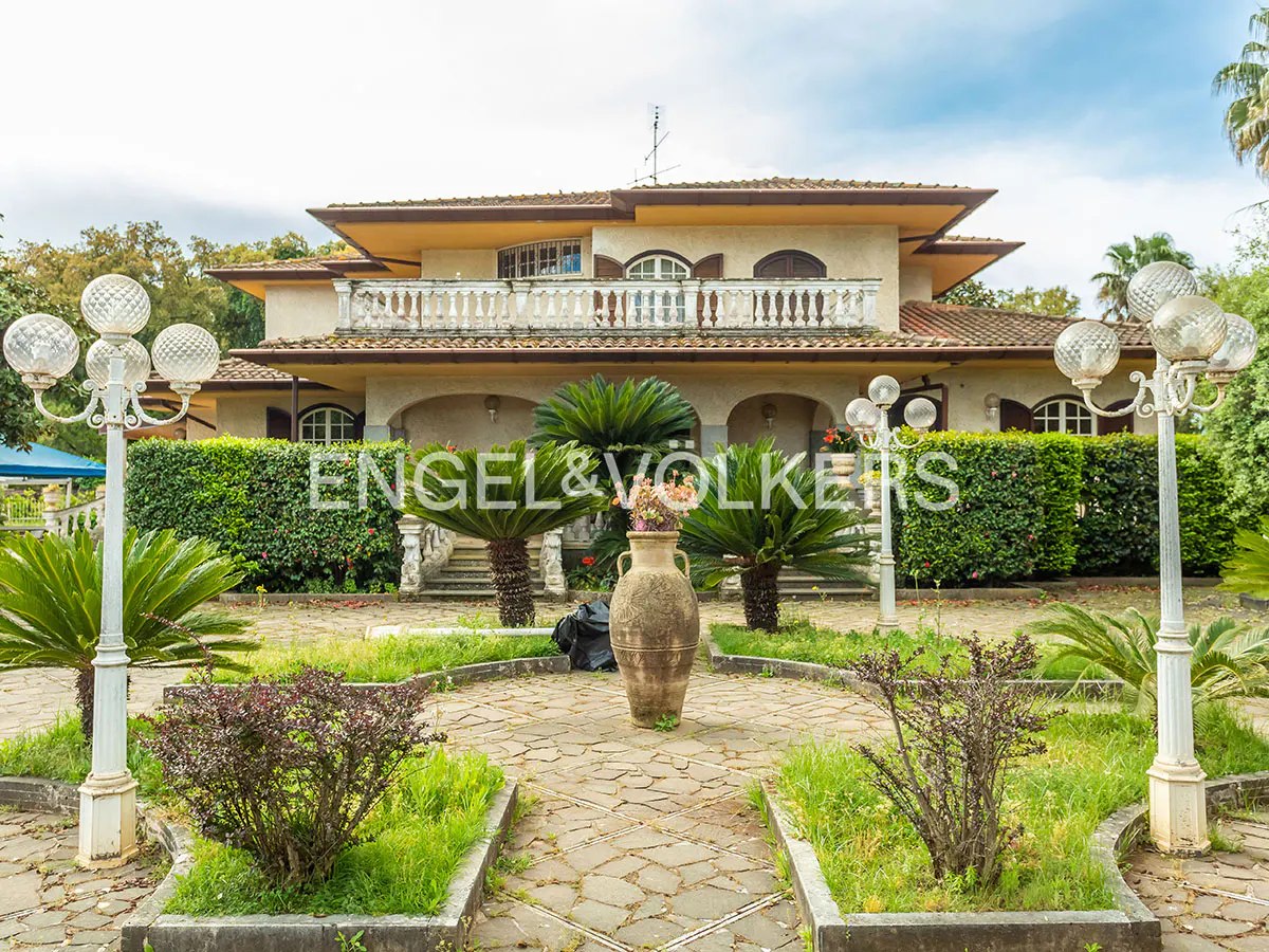 Two-story beige house with a balcony, surrounded by a garden with palm trees and a large vase.