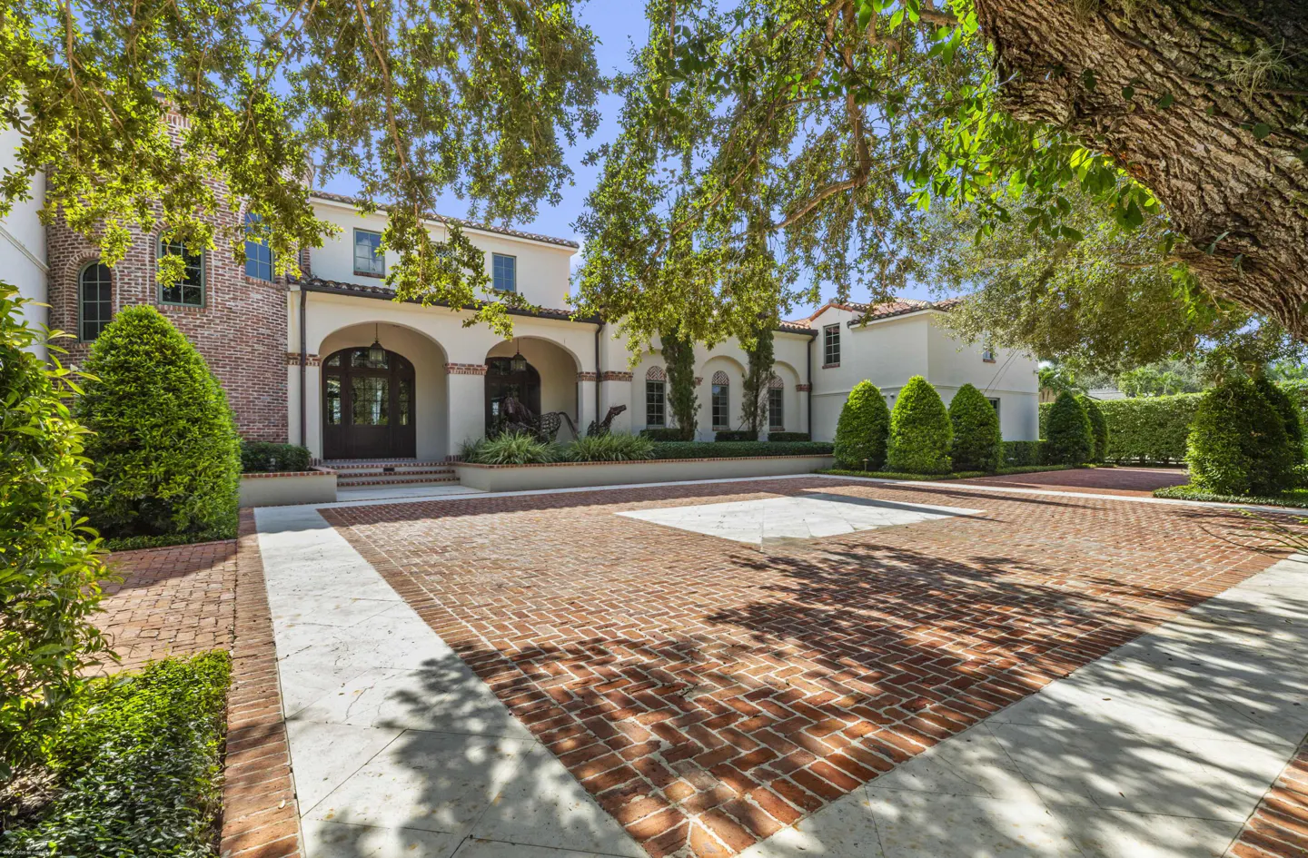 Exterior view of a two-story white house with a brick driveway and arched entryway. Green trees frame the house.