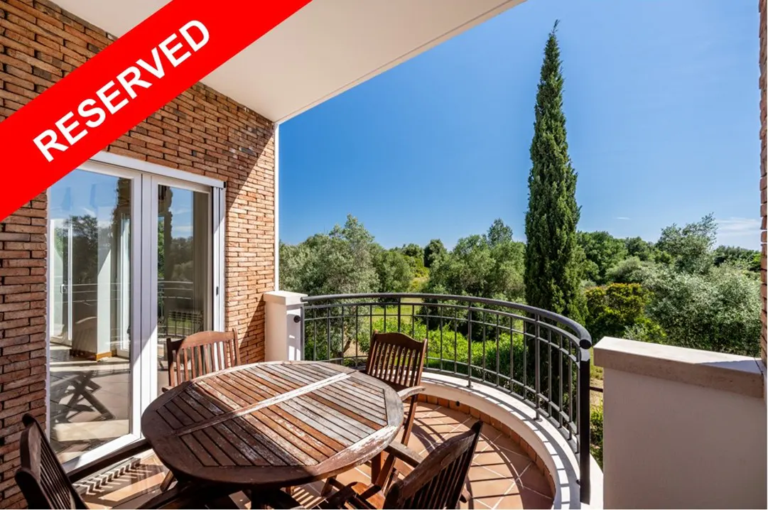 Reserved balcony view with a round wooden table, chairs, and black railing overlooking green trees under a blue sky.