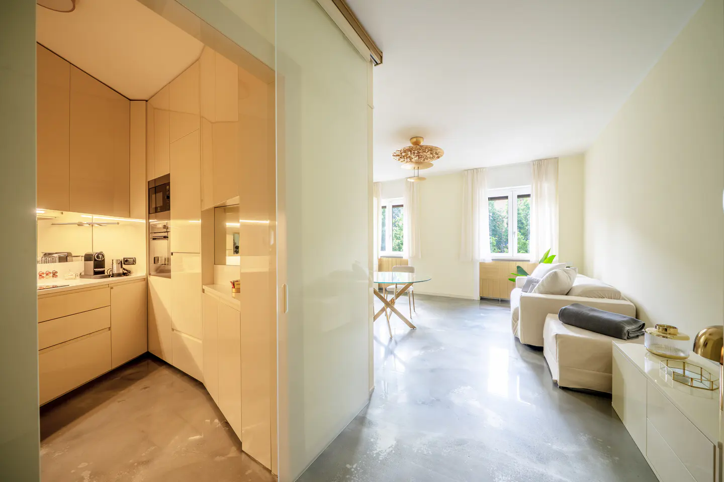 Open-concept apartment with a kitchen featuring glossy beige cabinets, and a living room with a white sofa and glass table.