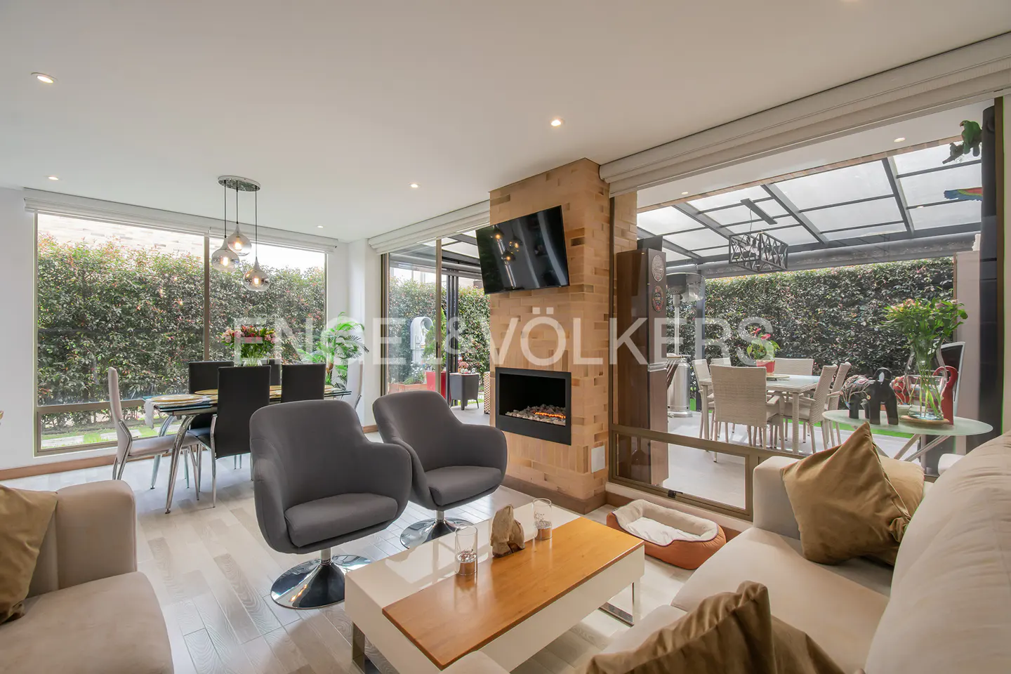 Bright living room with gray chairs, a brick fireplace with a TV, and a dining area visible through a large window.