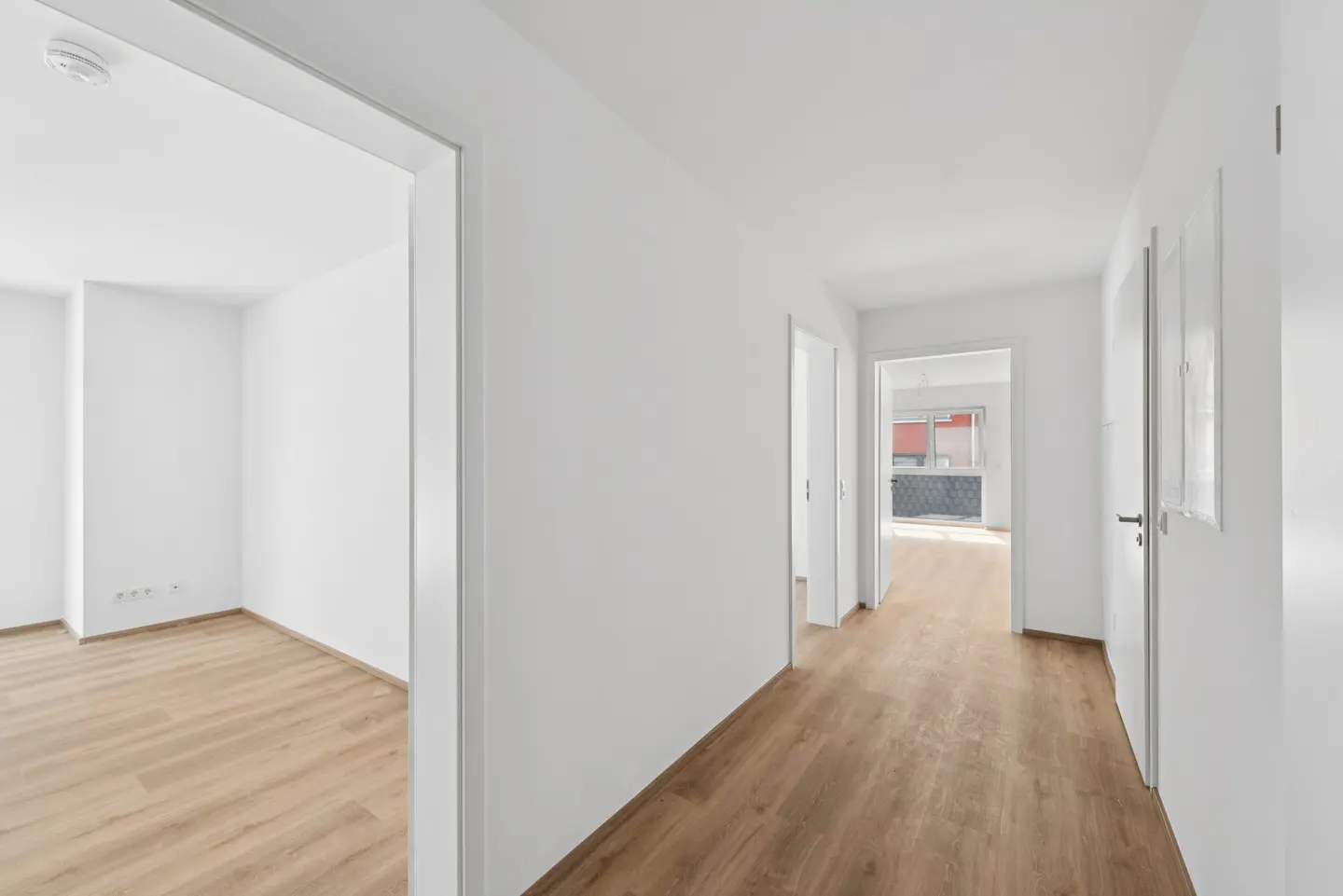 Bright, empty hallway with light wood floors and white walls. Open doorway to a room on the left. Doors line the hallway on the right.