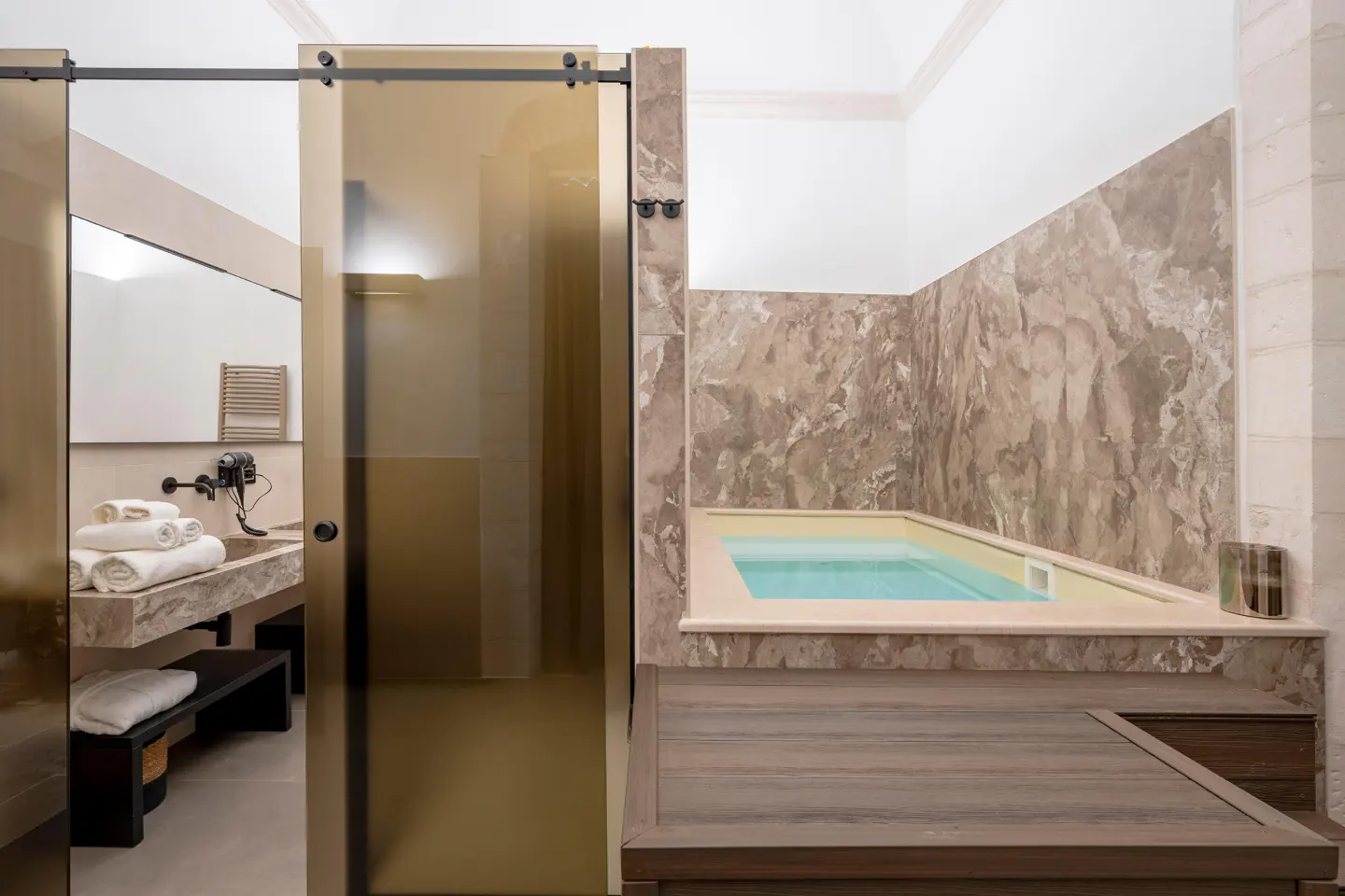 Bathroom with a marble-walled jacuzzi and a sliding glass door revealing a sink with stacked white towels.