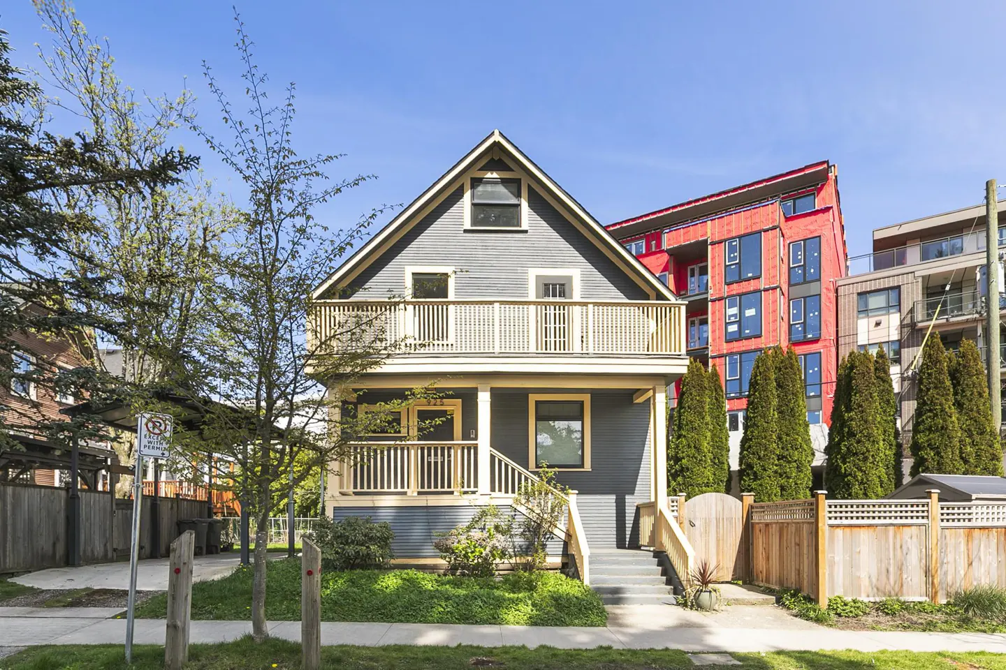 Two-story gray house with a cream-colored balcony and trim, set against a blue sky and green trees. A red building is visible in the background.
