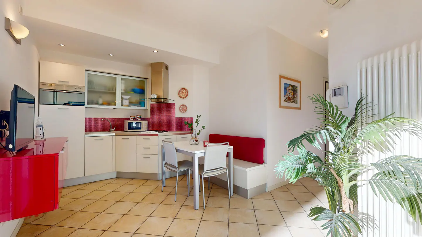 A bright kitchen and dining area with white cabinets, red backsplash, and a white table with chairs. A red cabinet and a green plant add color.