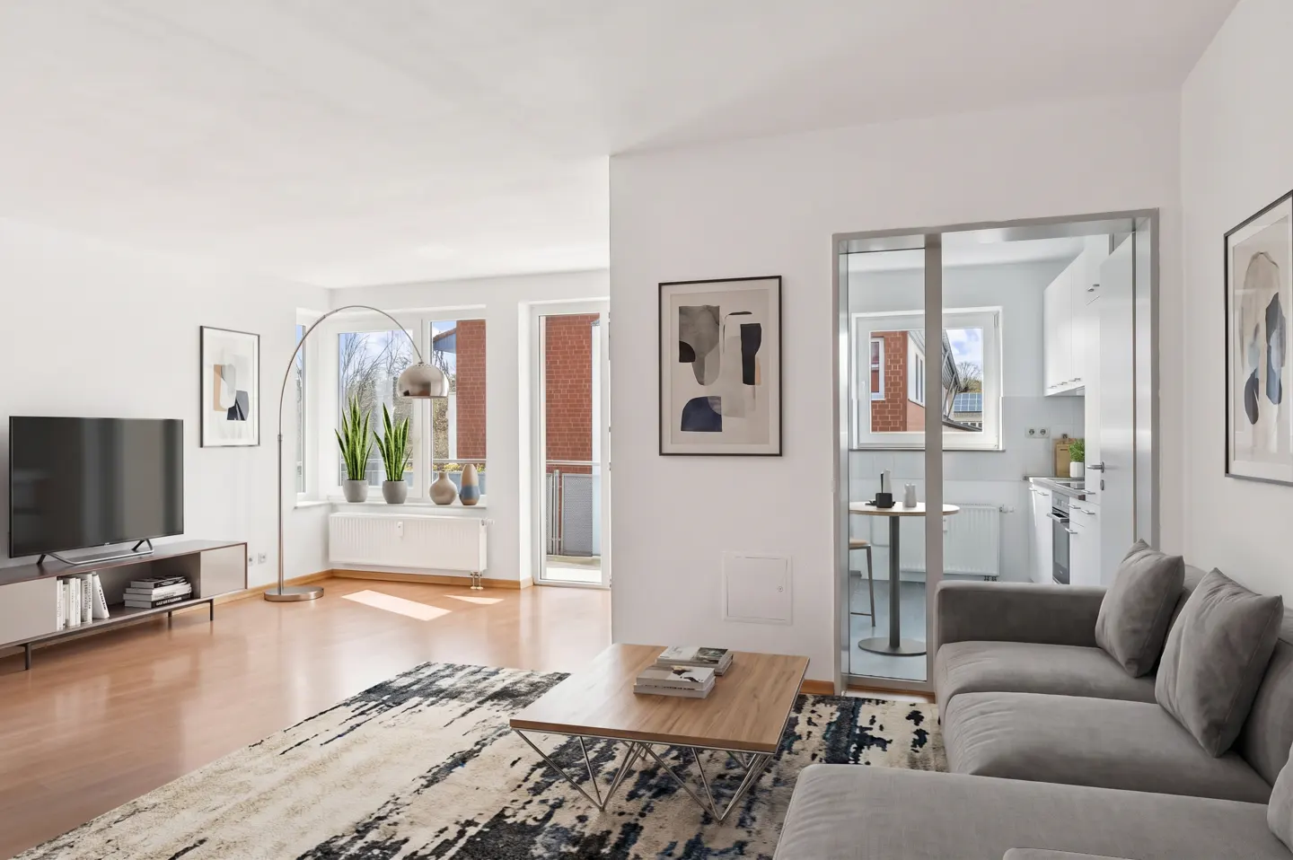 Bright living room with wood floors, white walls, gray sofa, TV, and a view of the kitchen through a glass door.