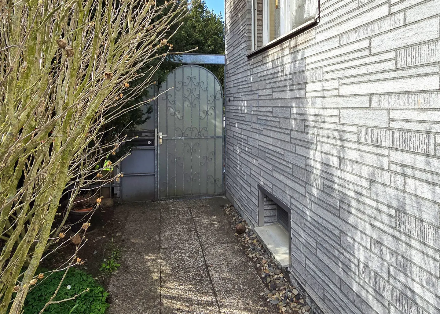 Side view of a gray brick house with a metal gate and a bare tree in the foreground.