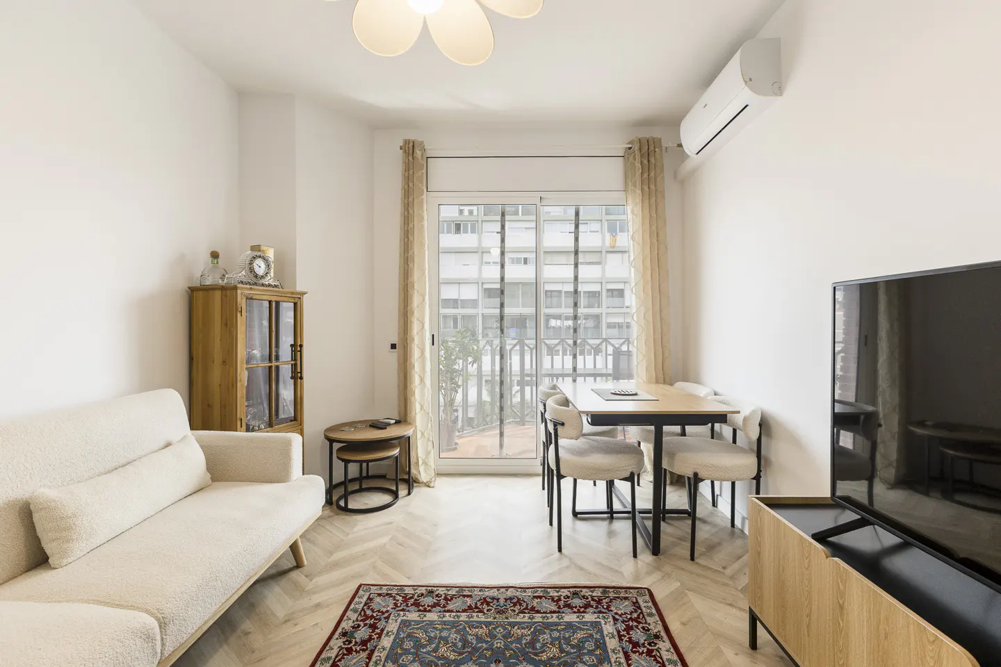 Bright living room with white walls, herringbone floors, and a blue patterned rug. A cream sofa, wood cabinet, and dining table with chairs furnish the space. Balcony doors let in natural light.