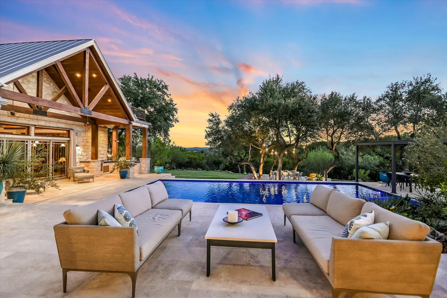 Outdoor patio with sofas, table, and pool at dusk. Stone house with wood beams in the background. Trees and sunset sky.