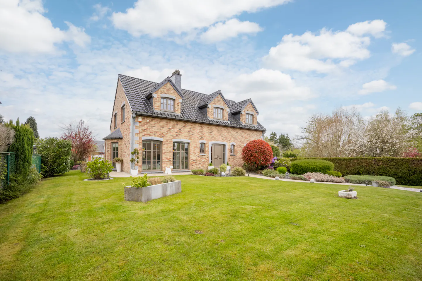 A tan brick house with a black roof sits on a large green lawn under a blue sky with white clouds.