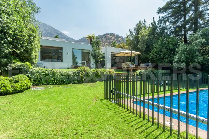 Exterior of a modern white house with a green lawn, pool with a black fence, trees, and a mountain in the background.