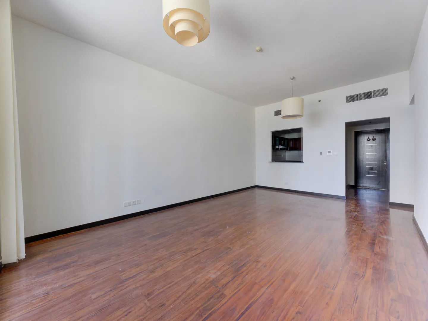 Empty living room with wood floors, white walls, and two hanging light fixtures. A doorway and kitchen pass-through are visible in the background.