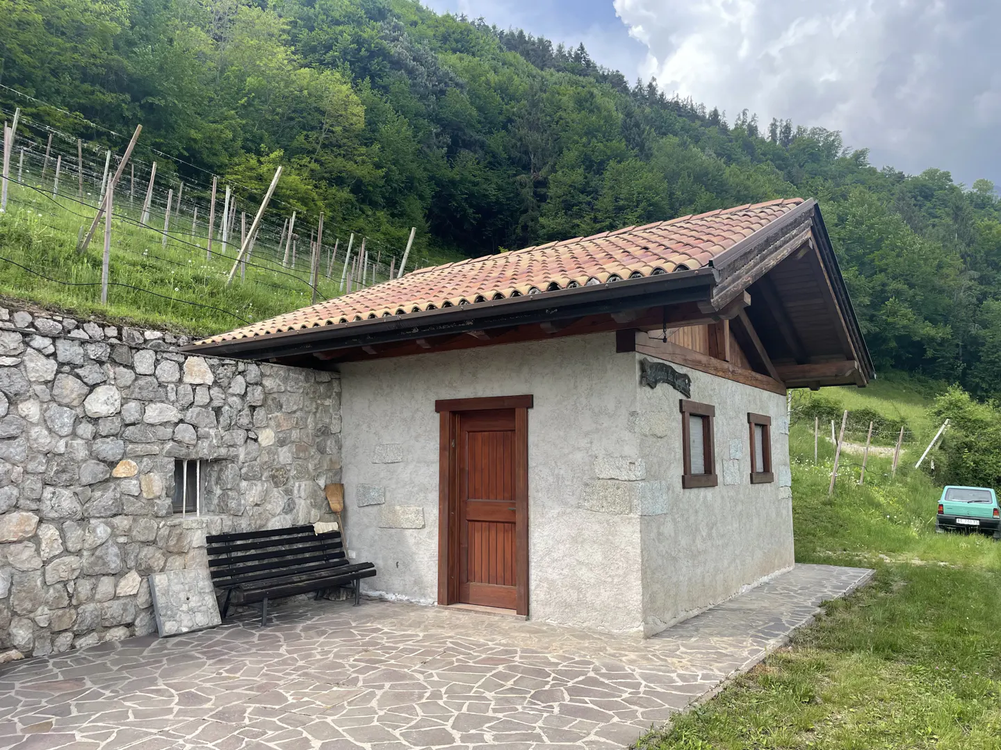 Stone building with a red tile roof, a wooden door, and a bench, set against a green hillside.