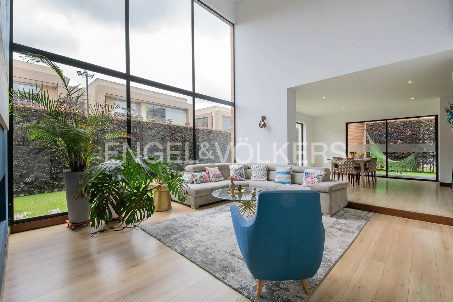 Modern living room with floor-to-ceiling windows, a grey sectional sofa, blue armchair, and a view of a green garden.