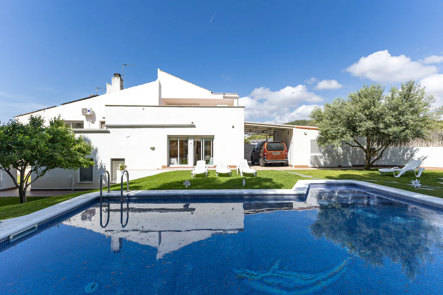A bright, white house with a blue-tiled pool in the foreground, surrounded by green grass and trees under a blue sky.