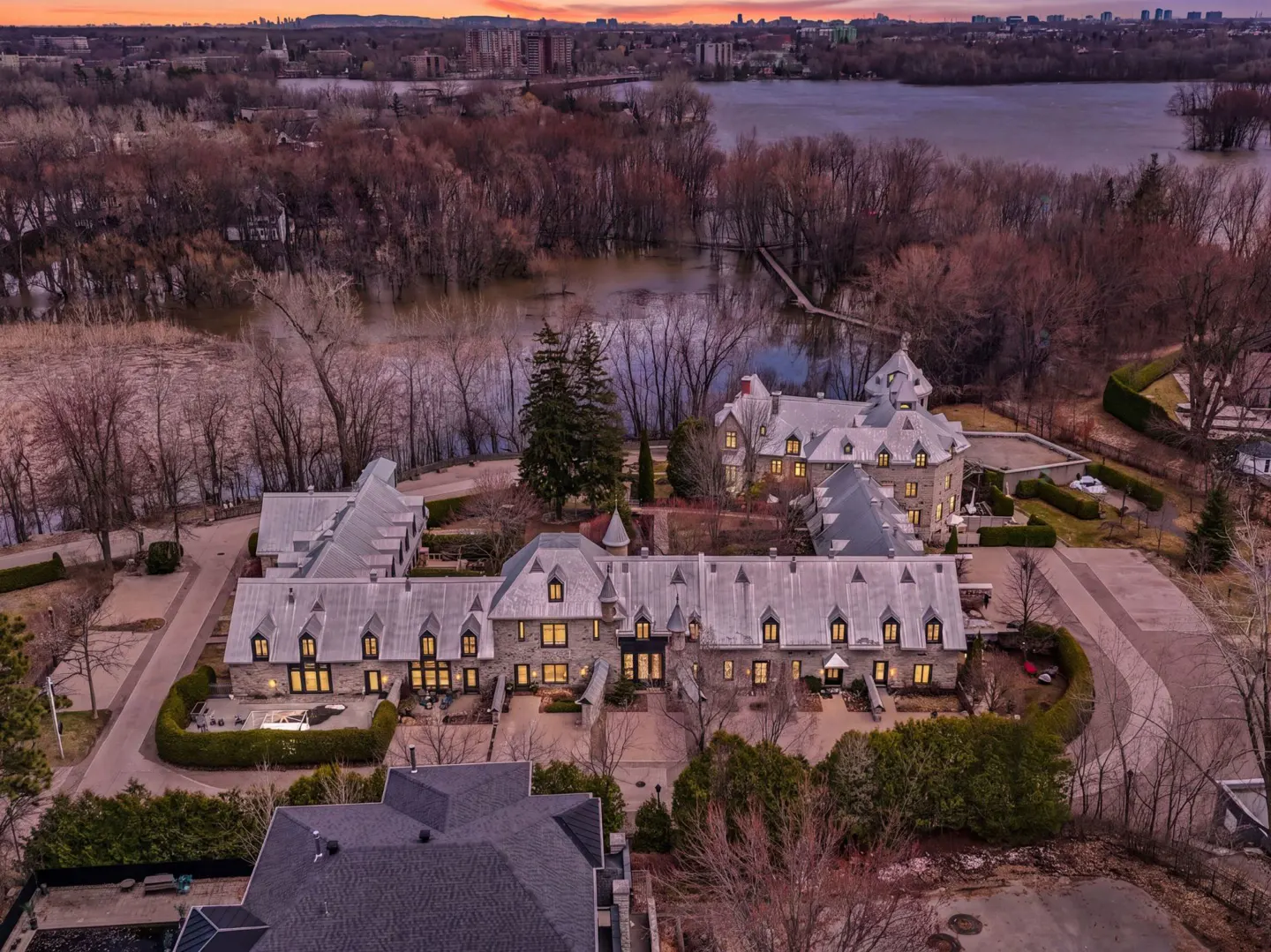 Aerial view of a large, stone mansion with grey roofs, surrounded by trees and water at sunset.