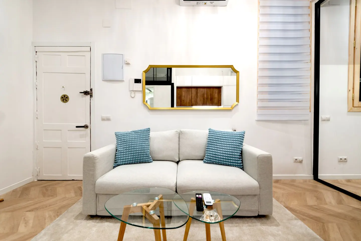 Living room with a white sofa, blue pillows, and a glass coffee table on a beige rug. A gold-framed mirror hangs above the sofa.