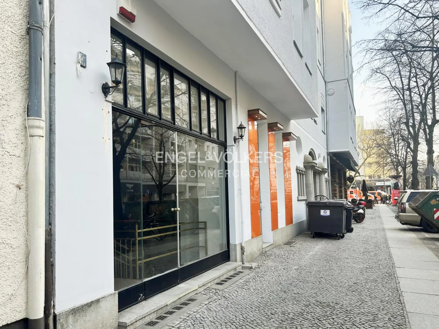 Exterior of a white building with a black-framed glass storefront for Engel & Volkers Commercial. Street view with bins and parked vehicles.