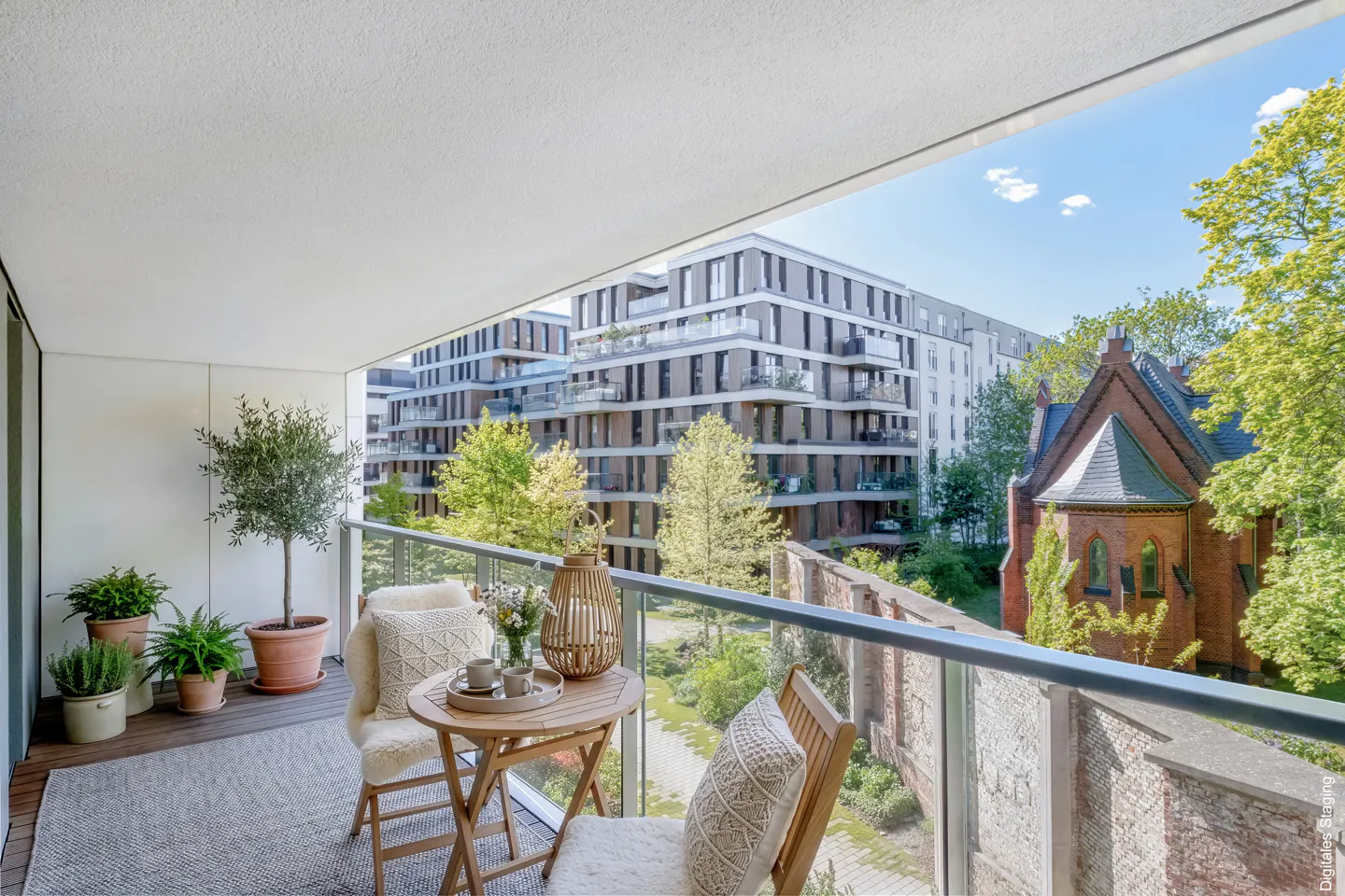 Balcony with a table, chairs, and plants overlooking a courtyard with buildings and trees.