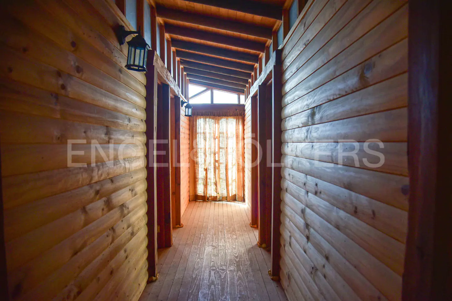 Hallway with wood-paneled walls and ceiling, leading to a window with sheer curtains. Lantern sconces on the walls.