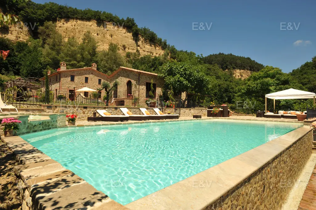 Exterior view of a stone house with a pool, lounge chairs, and a gazebo surrounded by trees.