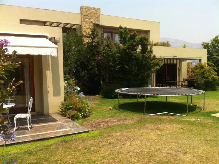Backyard view of a tan house with a trampoline on the green lawn. Patio with white furniture and flowers.