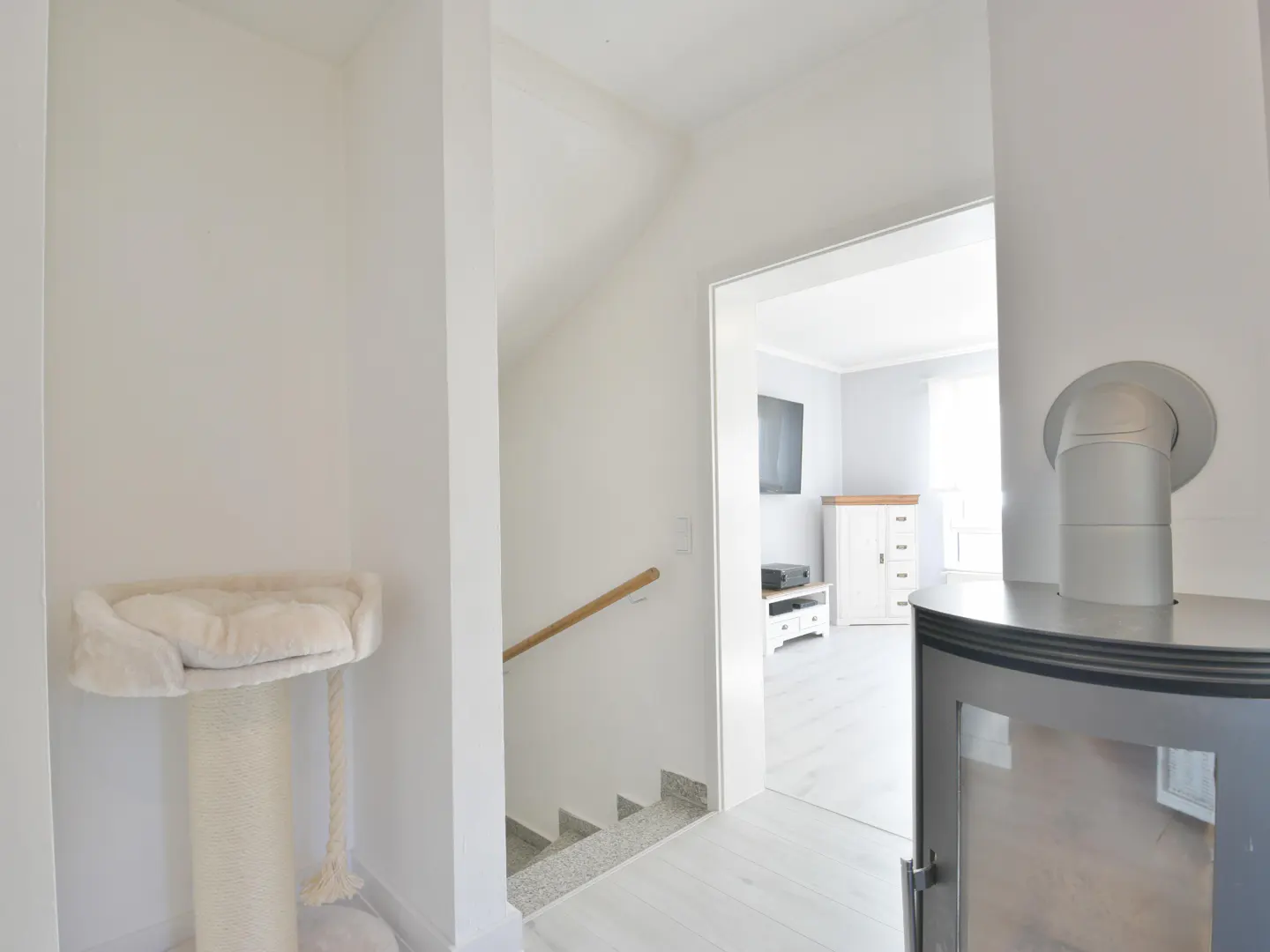 Bright, white interior with a cat tree, stairs, and a glimpse into a living room with a TV and cabinet.