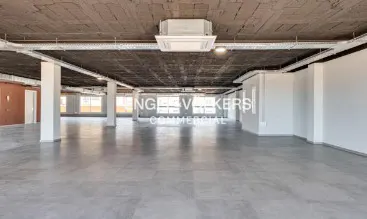 Empty commercial space with gray floors, white walls, and exposed ceiling. Columns support the ceiling. "Engel & Volkers Commercial" is visible in the distance.