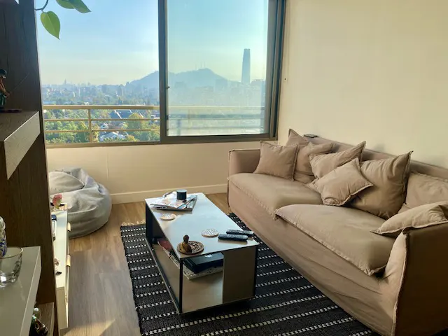 Living room with a beige sofa, pillows, coffee table, and a view of a city skyline through a large window.