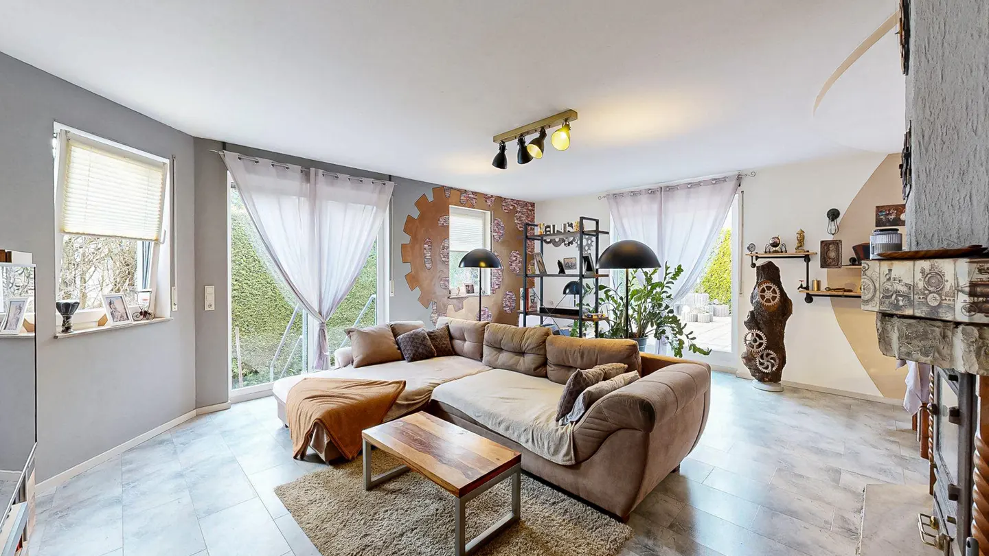 Living room with brown sectional sofa, wood coffee table, and gear-themed decor. Windows with sheer curtains let in natural light.