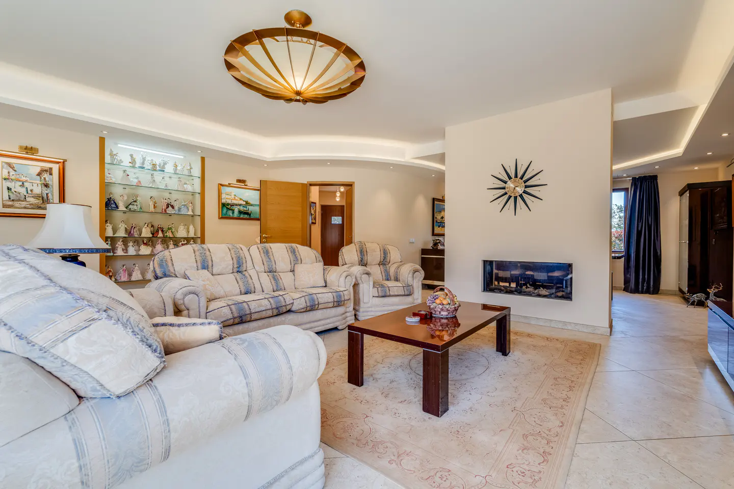 Living room with sofas, a coffee table, and a fireplace. A display cabinet with figurines is on the left. A starburst clock hangs above the fireplace.
