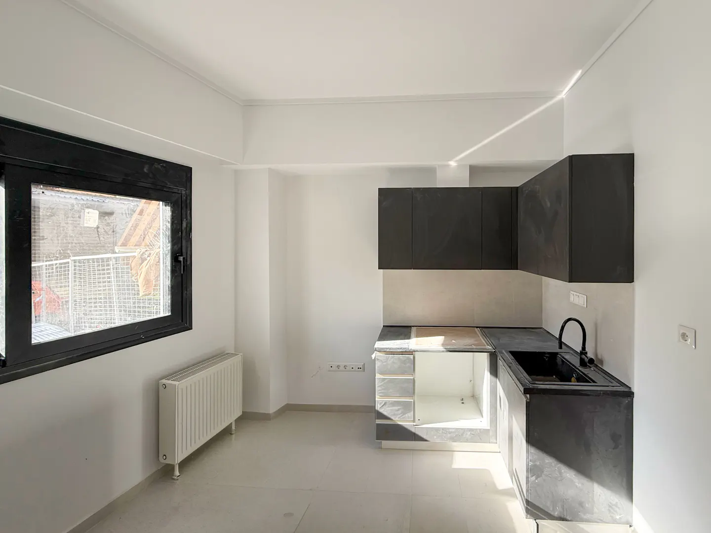 Bright, empty kitchen with white walls, gray cabinets, and a black sink. A black-framed window is on the left, next to a white radiator.