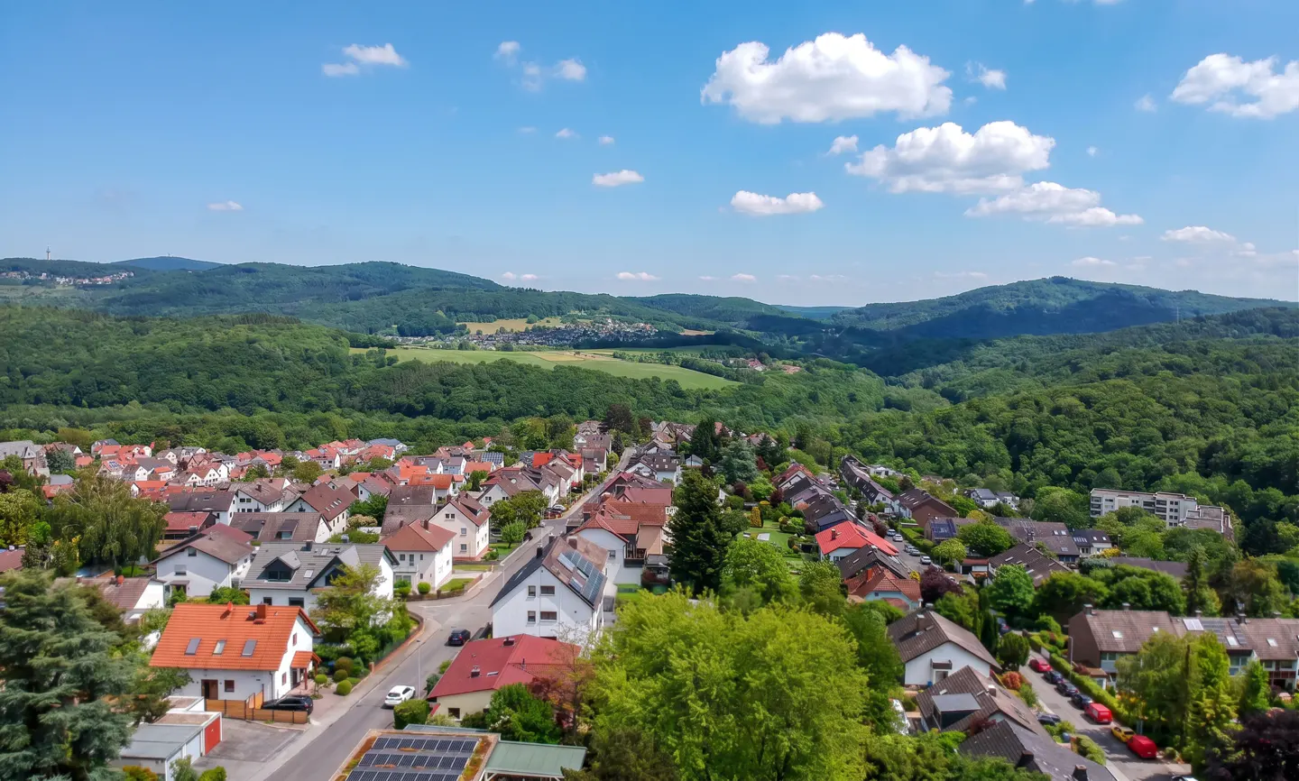 Aerial view of a residential neighborhood with houses featuring red roofs, surrounded by lush green forests and rolling hills.