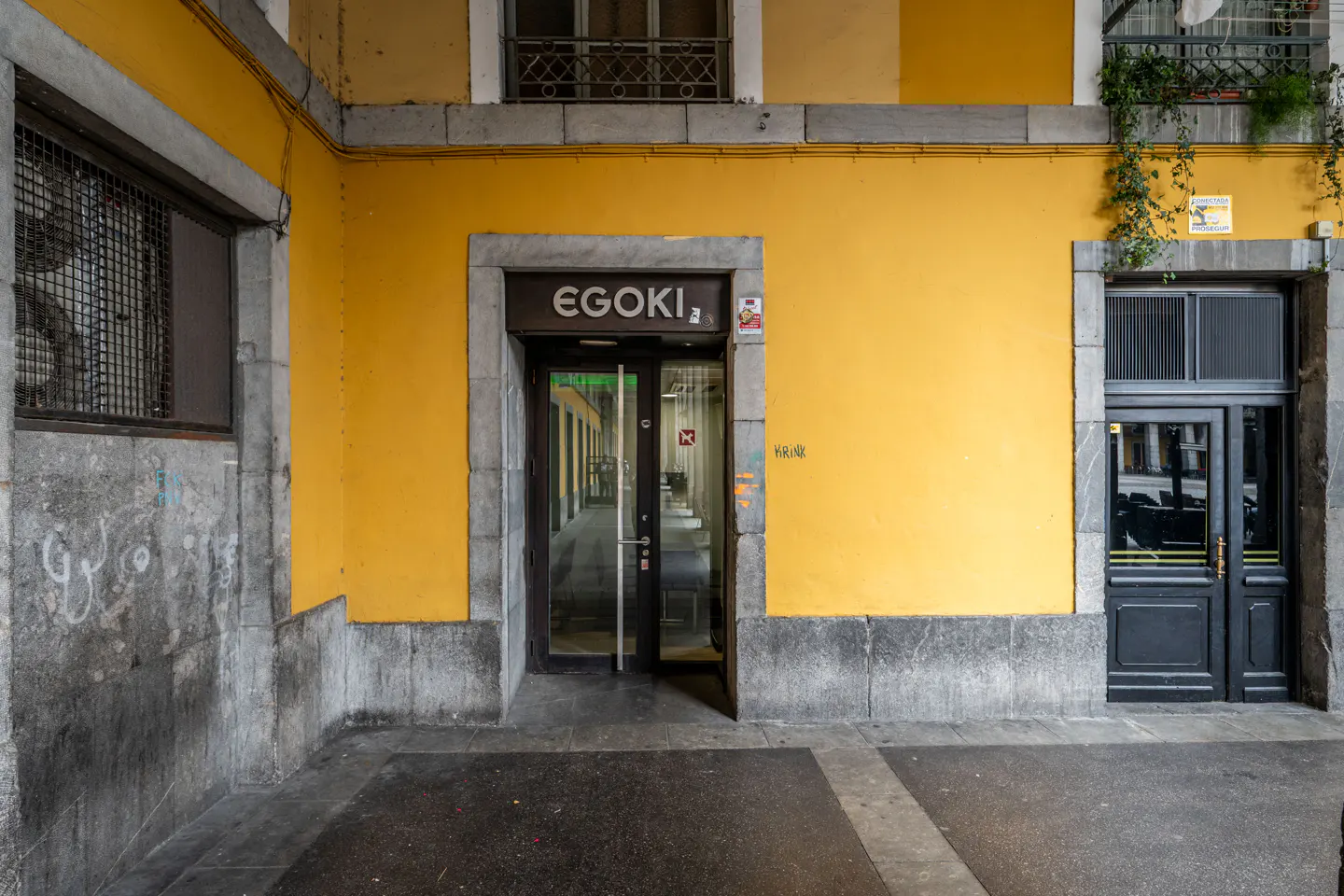 Entrance to Egoki with a glass door, set in a bright yellow building facade with grey stone trim and a dark side door.