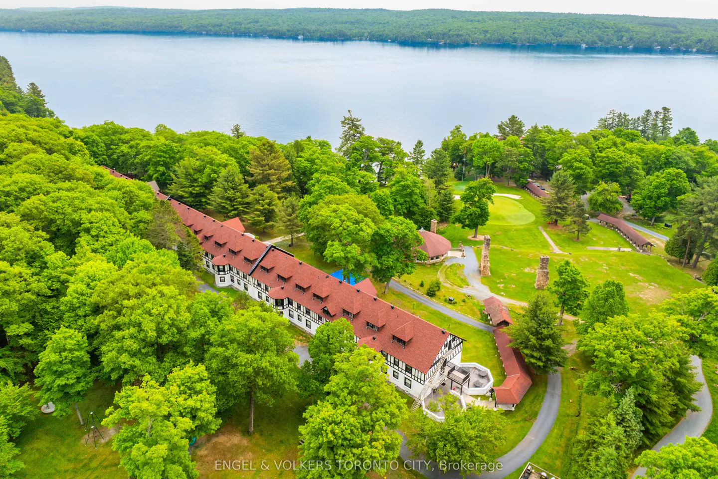 Aerial view of a long, white building with a red roof, surrounded by green trees and a lake in the background.