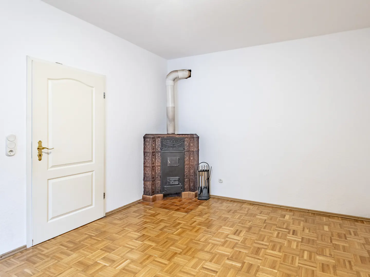 Bright room with parquet floor, white walls, and a tiled stove with a metal chimney. A white door is on the left.