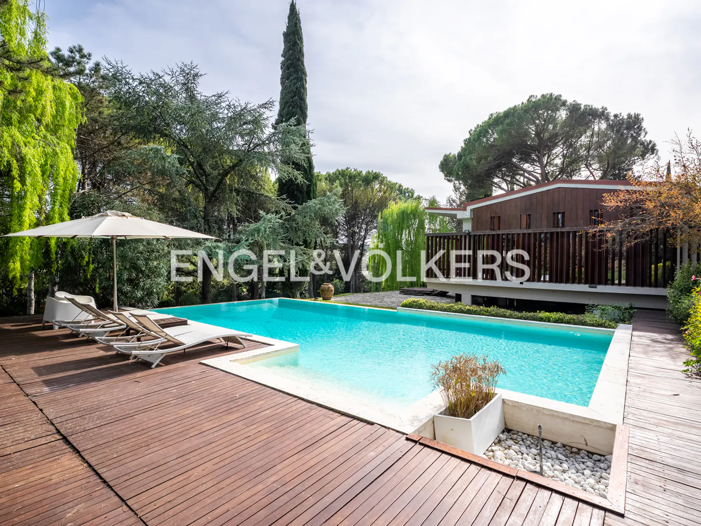 Exterior view of a pool with lounge chairs, umbrella, and wood deck, surrounded by trees, with a modern house in the background.