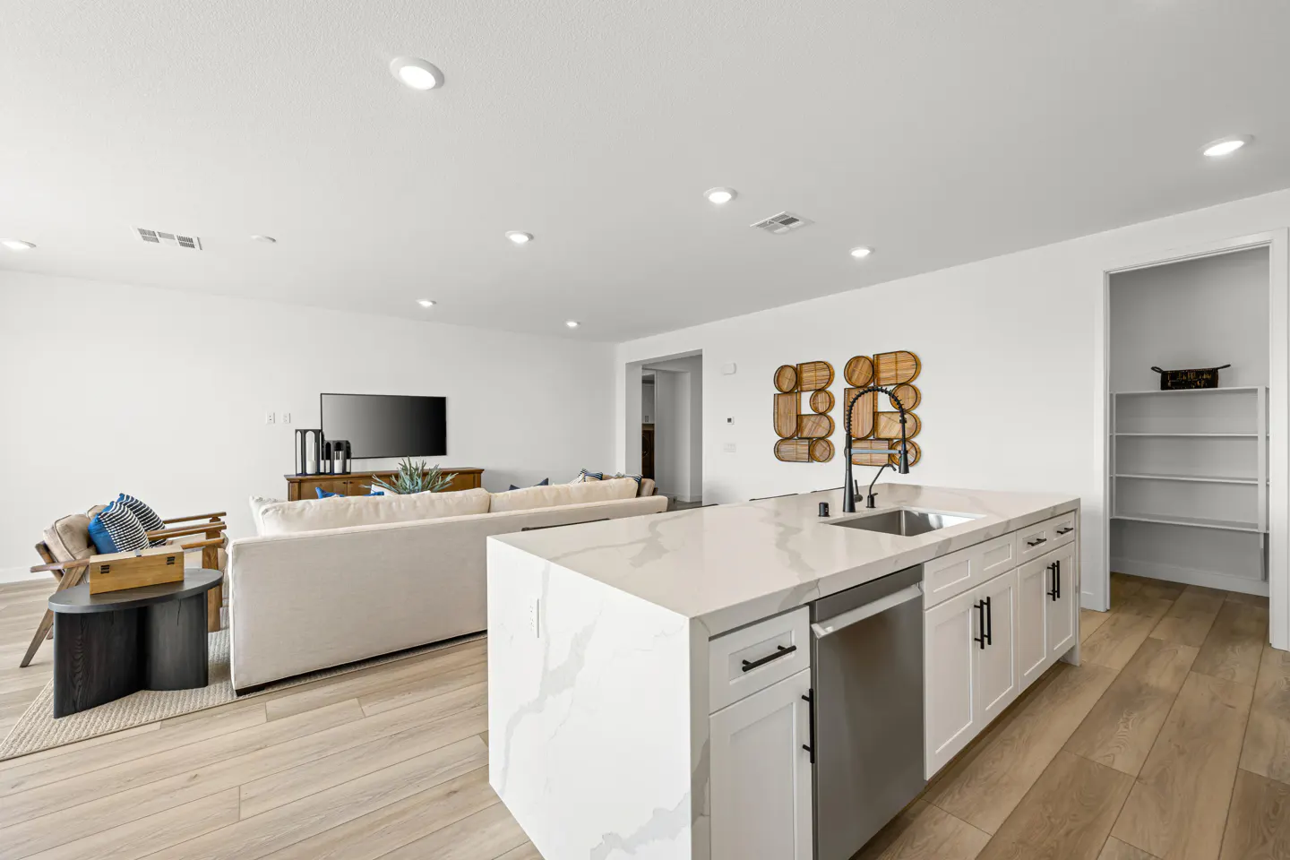 Bright, open-concept living space with white walls, light wood floors, and a kitchen island. A white sofa and TV are in the background.