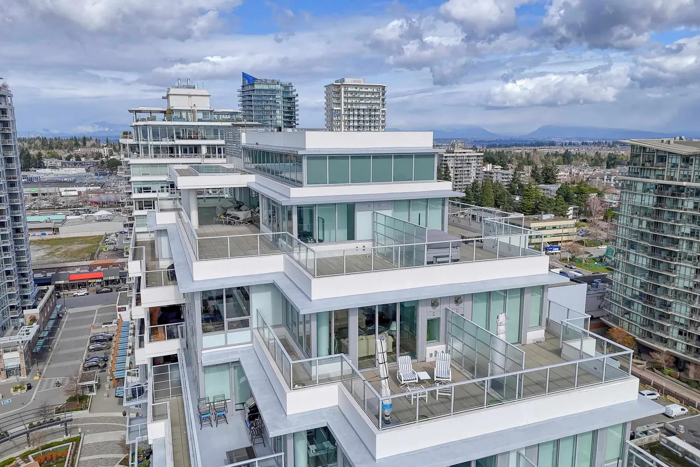 Aerial view of a modern white high-rise building with glass balconies and outdoor furniture, set against a cityscape and cloudy sky.