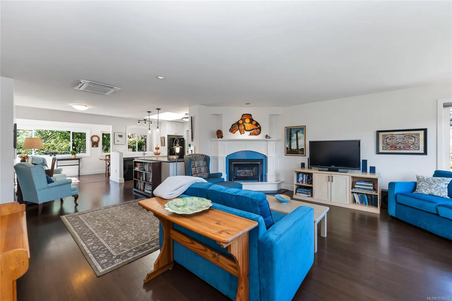 Bright, open-concept living room with blue sofas, a fireplace with a turtle art piece, and a kitchen in the background.