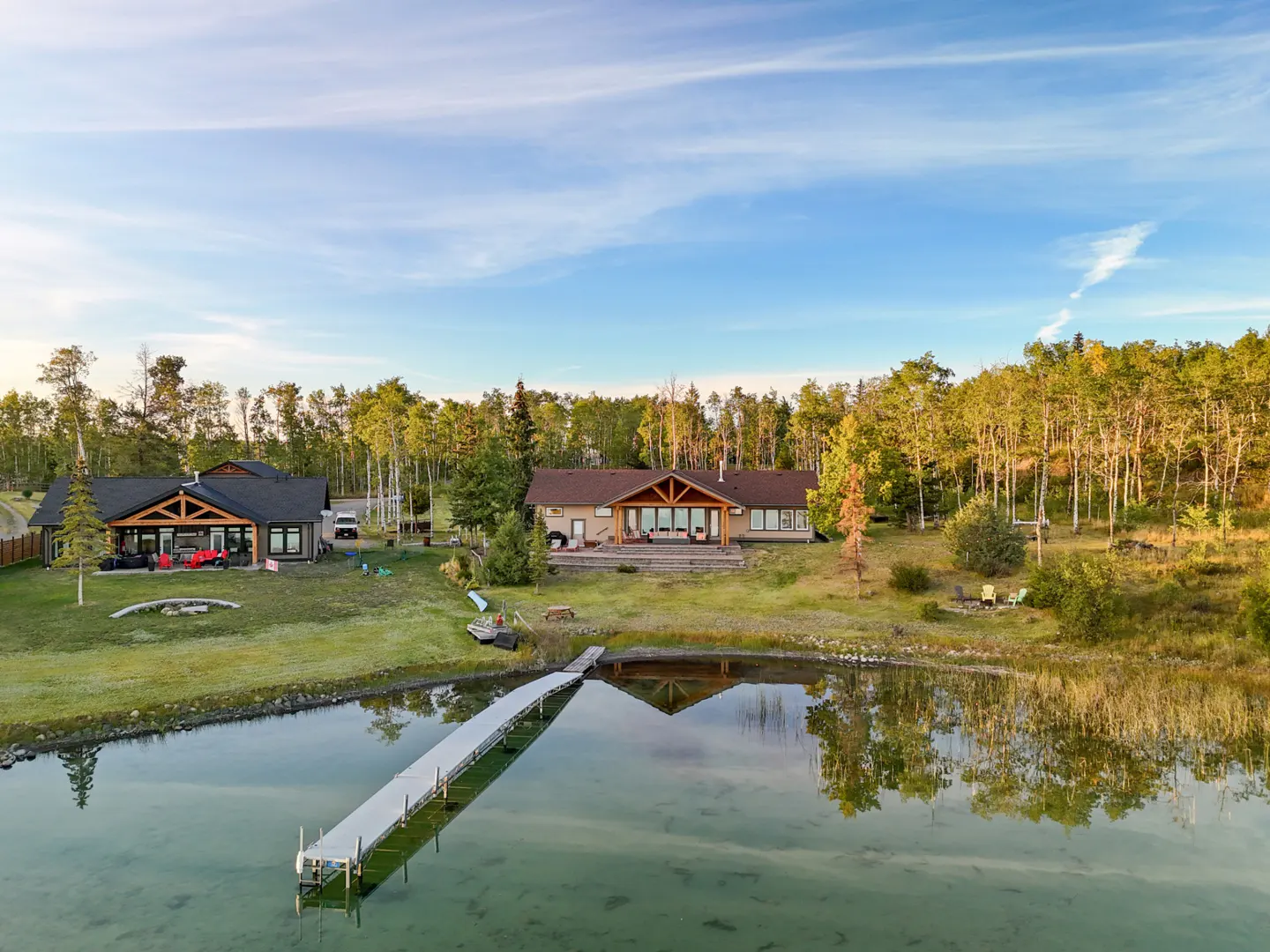 A lakeside property with two houses, a long dock, and a backdrop of trees under a blue sky.