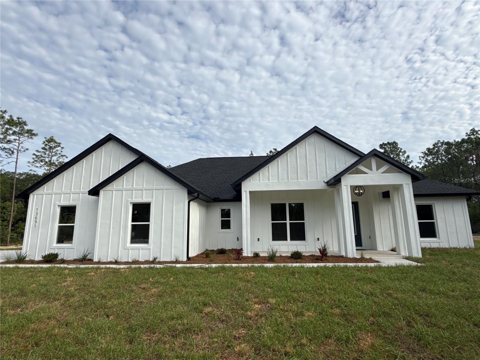 Modern white farmhouse with black roof and trim, featuring a covered porch and green lawn under a cloudy sky.