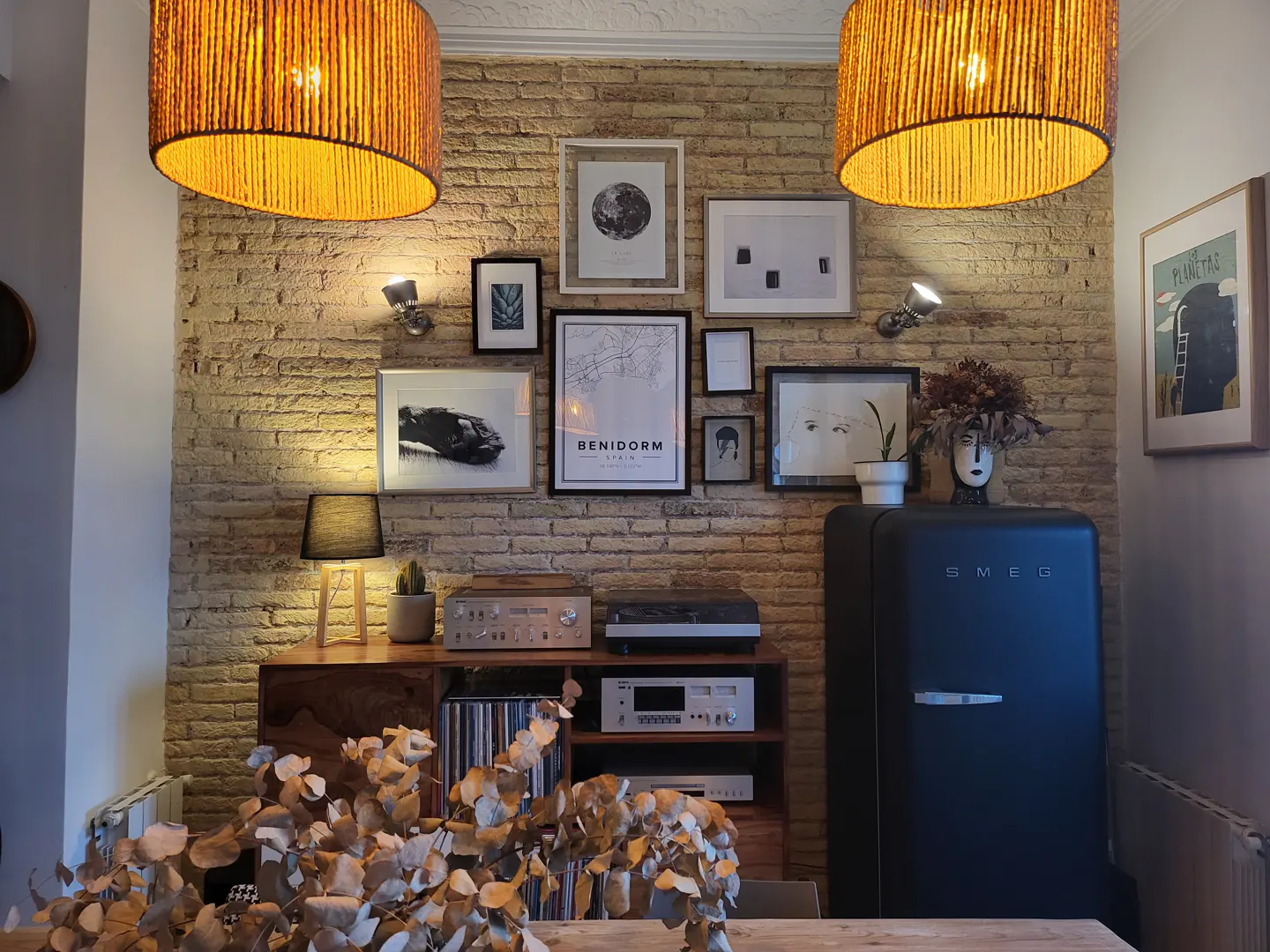 Cozy dining room with exposed brick wall, framed art, and warm lighting. A black SMEG refrigerator stands beside a wooden cabinet with vintage electronics.