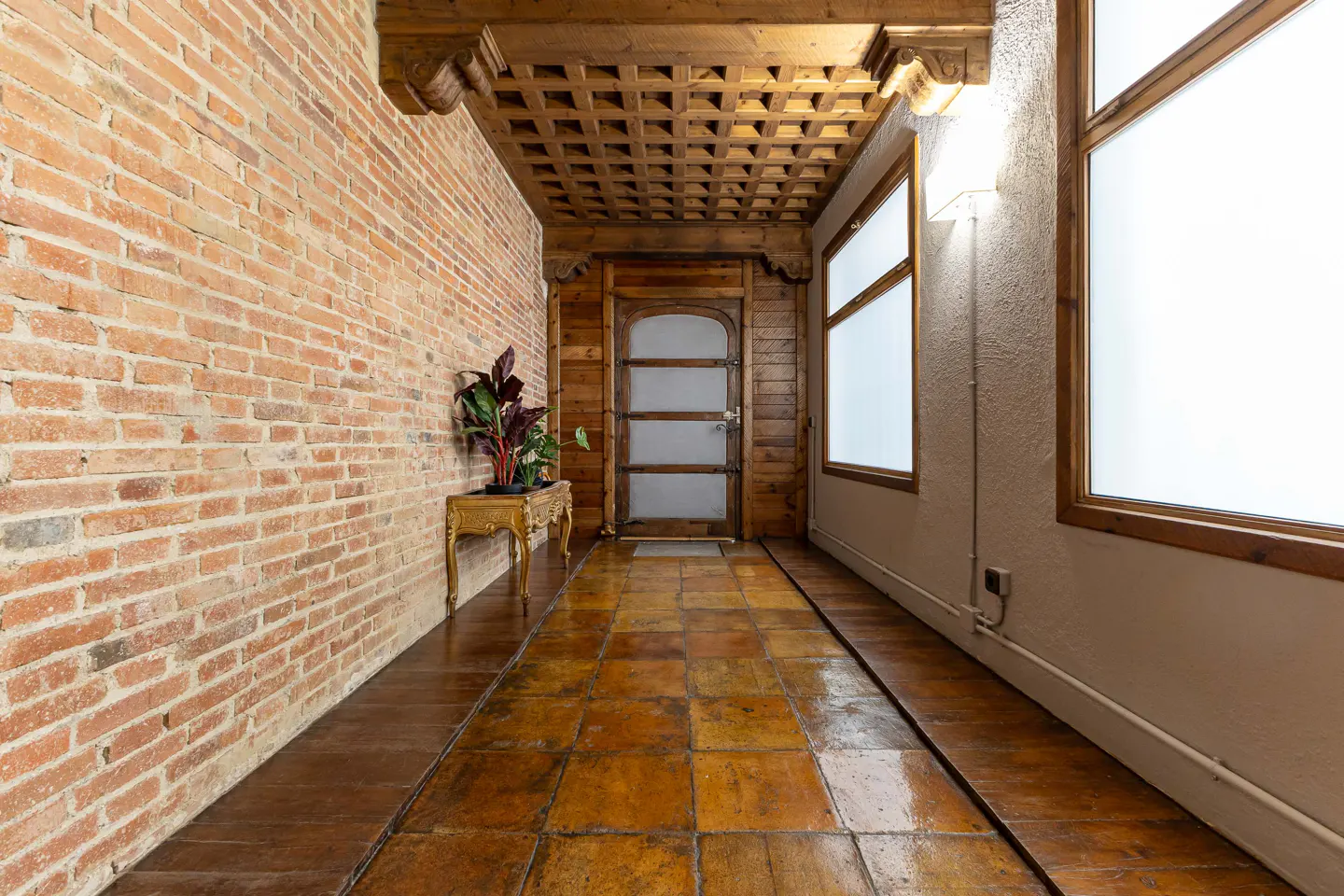 Hallway with brick wall, terracotta tile floor, wood ceiling, and frosted glass door. A plant sits on a gold table.