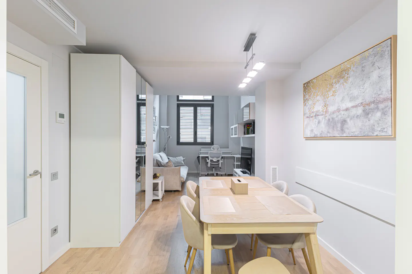 Bright, modern apartment interior with a light wood dining table and chairs. A living area with a sofa and desk is visible in the background.
