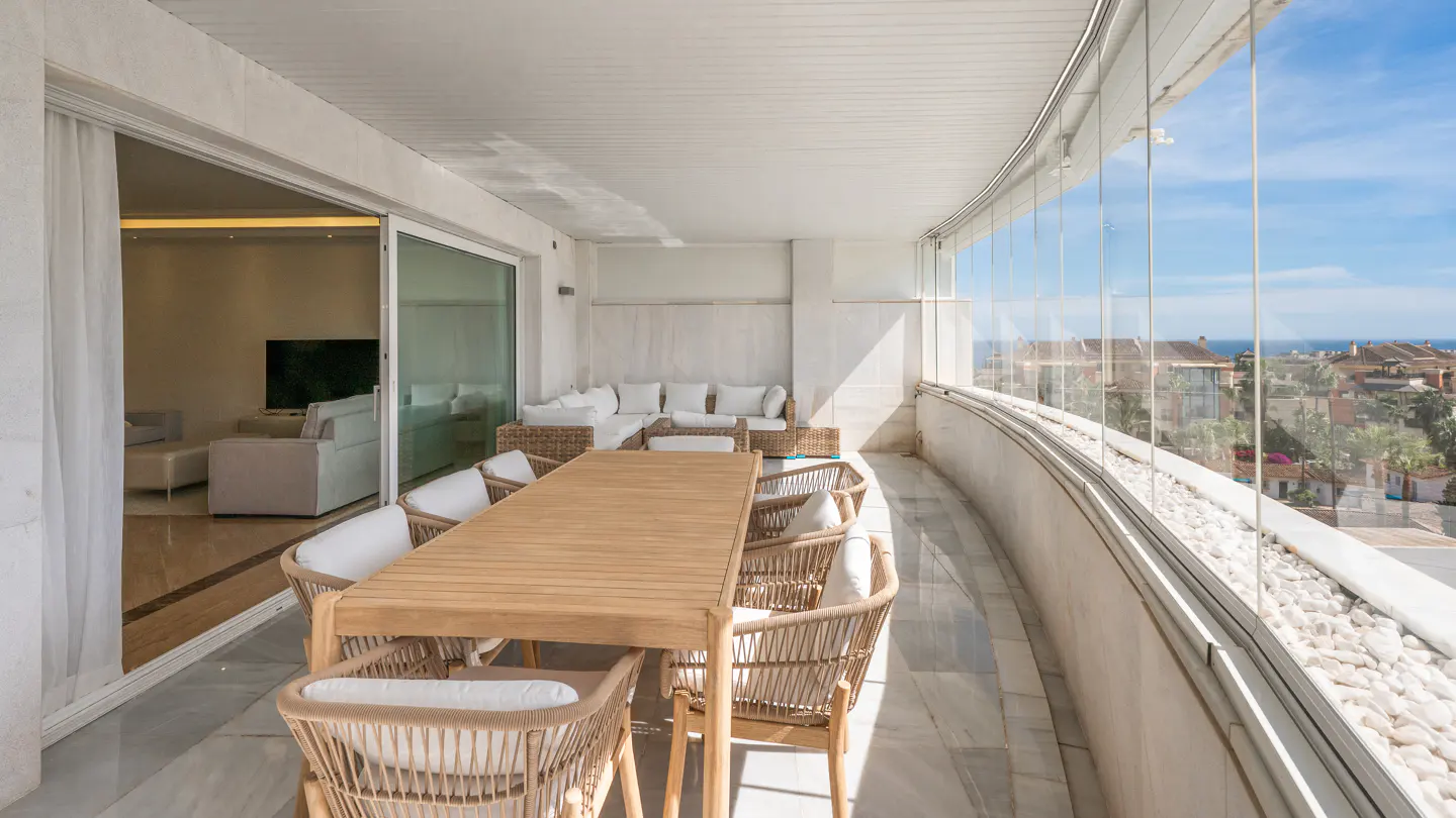Modern balcony with a wooden dining set, wicker chairs, and glass walls overlooking a sunny coastal neighborhood.